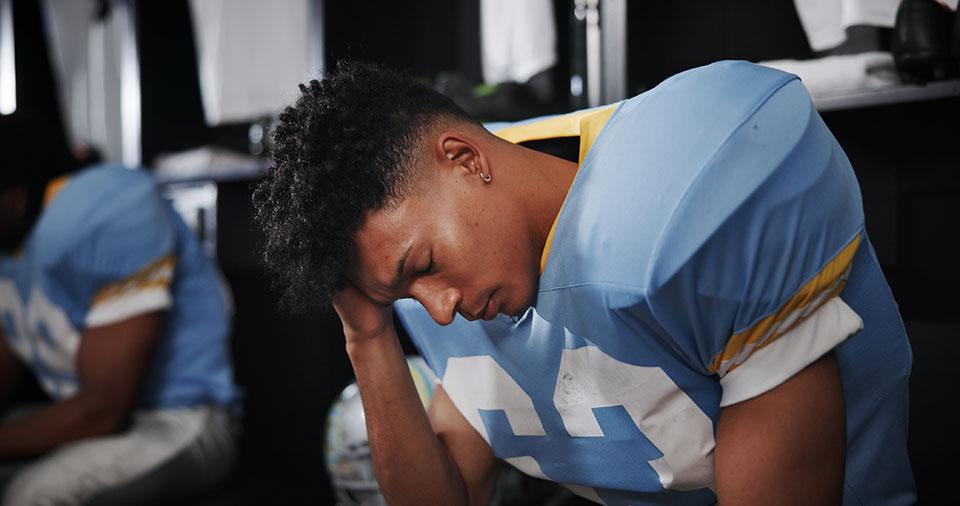 Male football player in a locker room after a sports concussion, holding his head in his hands.